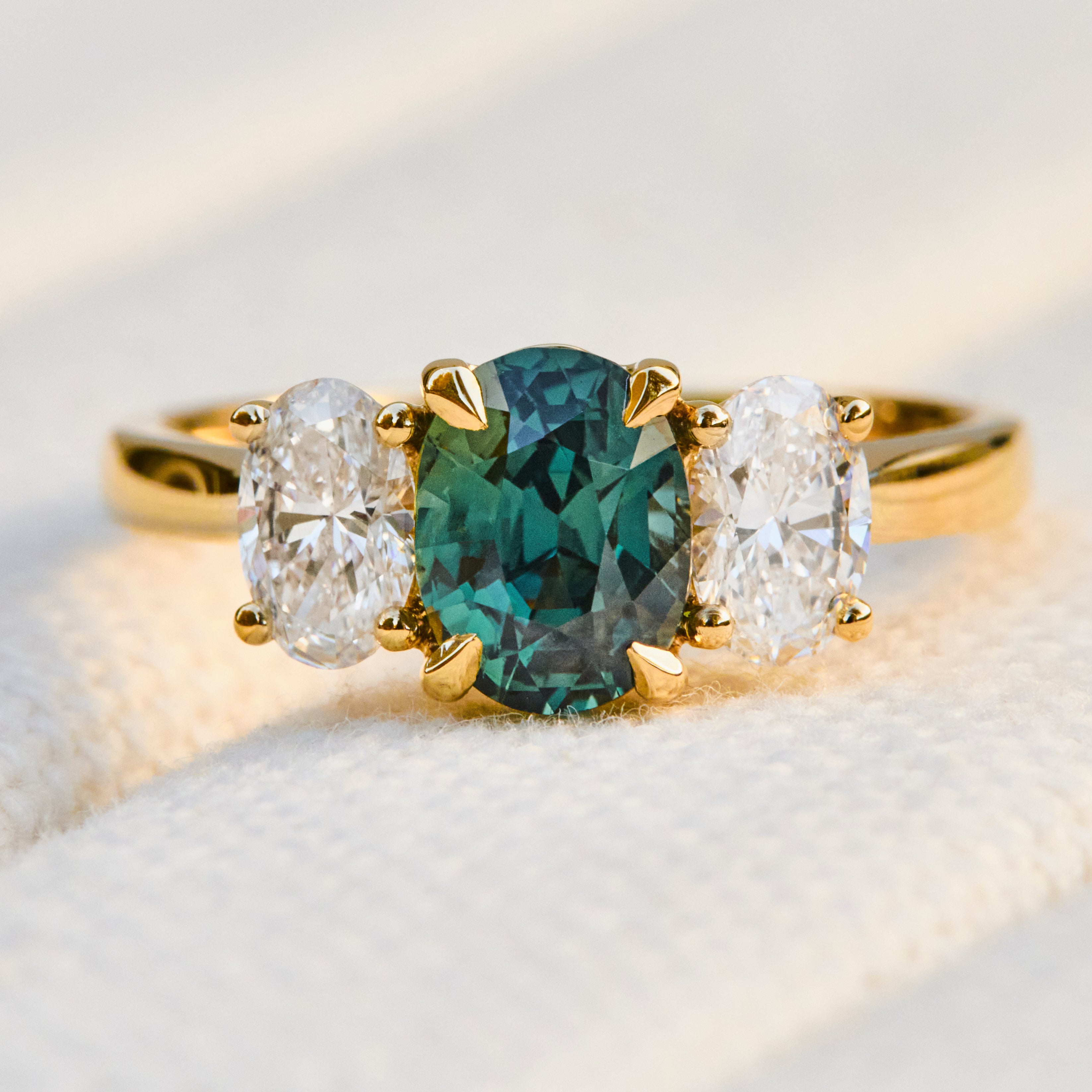 An 18ct yellow gold trilogy ring featuring a central oval teal sapphire flanked by two oval lab grown diamonds
