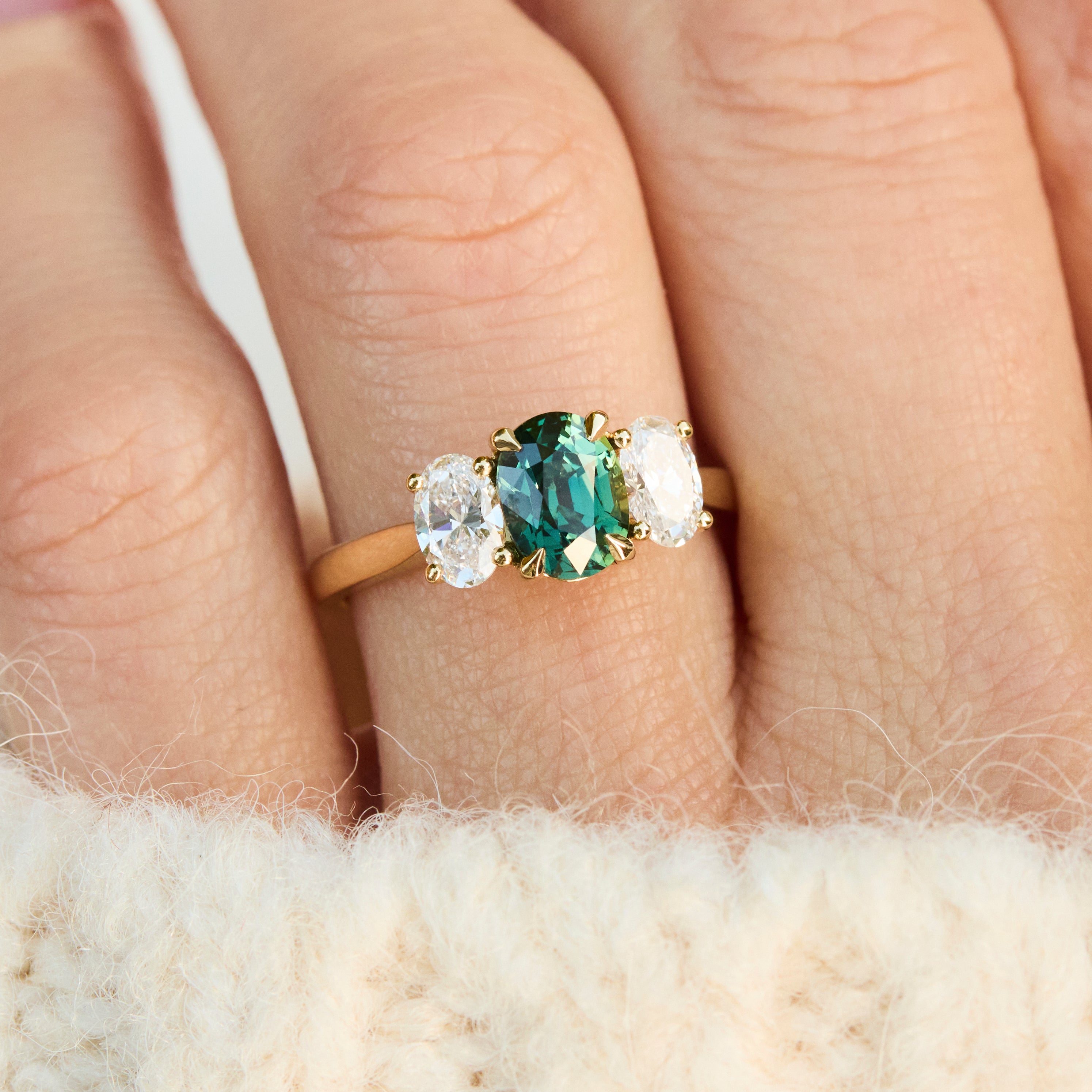A close-up of an 18ct yellow gold three-stone ring, featuring a central green oval sapphireflanked by two clear round-cut lab grown side stones.