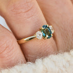 A close-up of an 18ct yellow gold engagement ring with a central oval teal sapphire stone accented by two small lab grown diamonds 