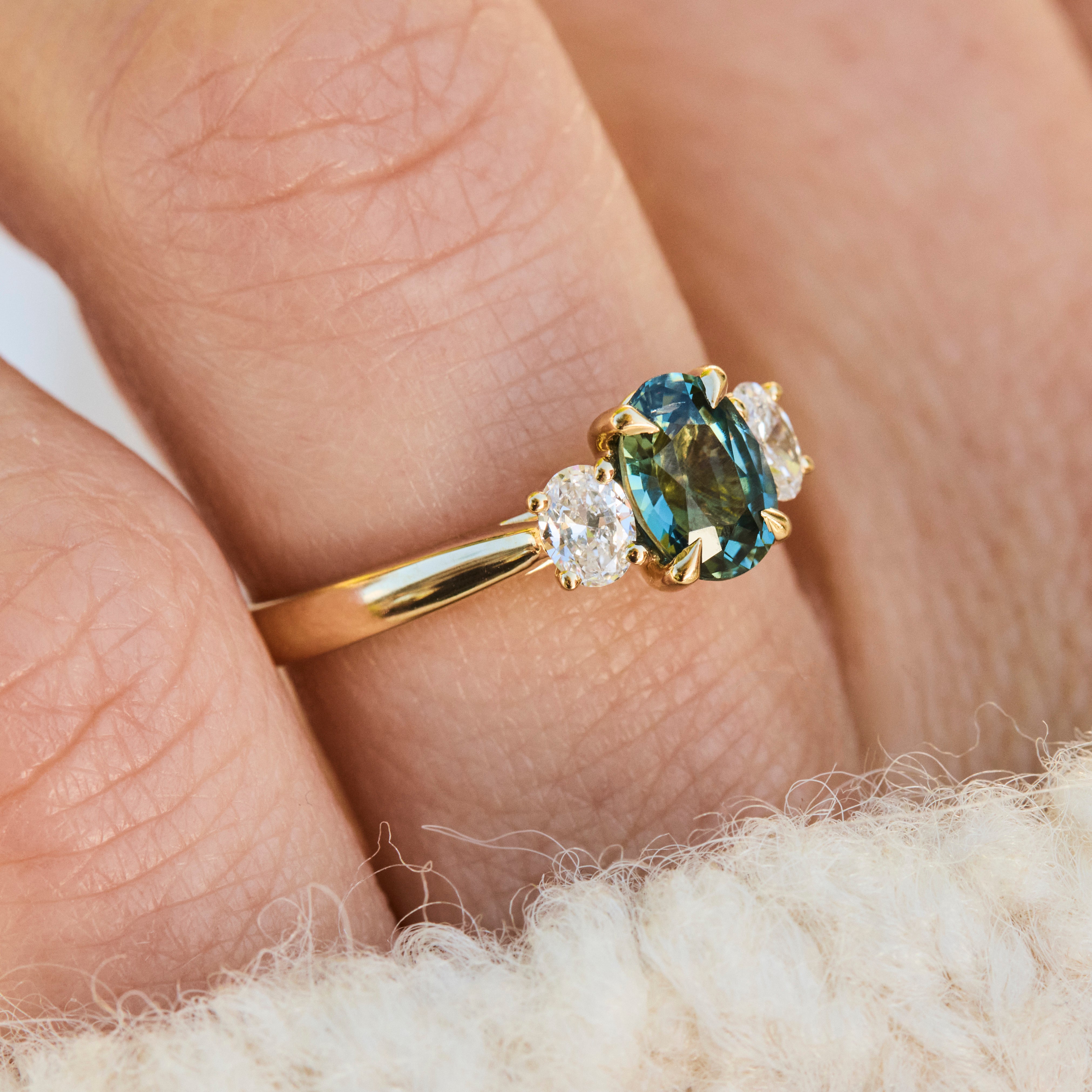 A close-up of an 18ct yellow gold engagement ring with a central oval teal sapphire stone accented by two small lab grown diamonds 