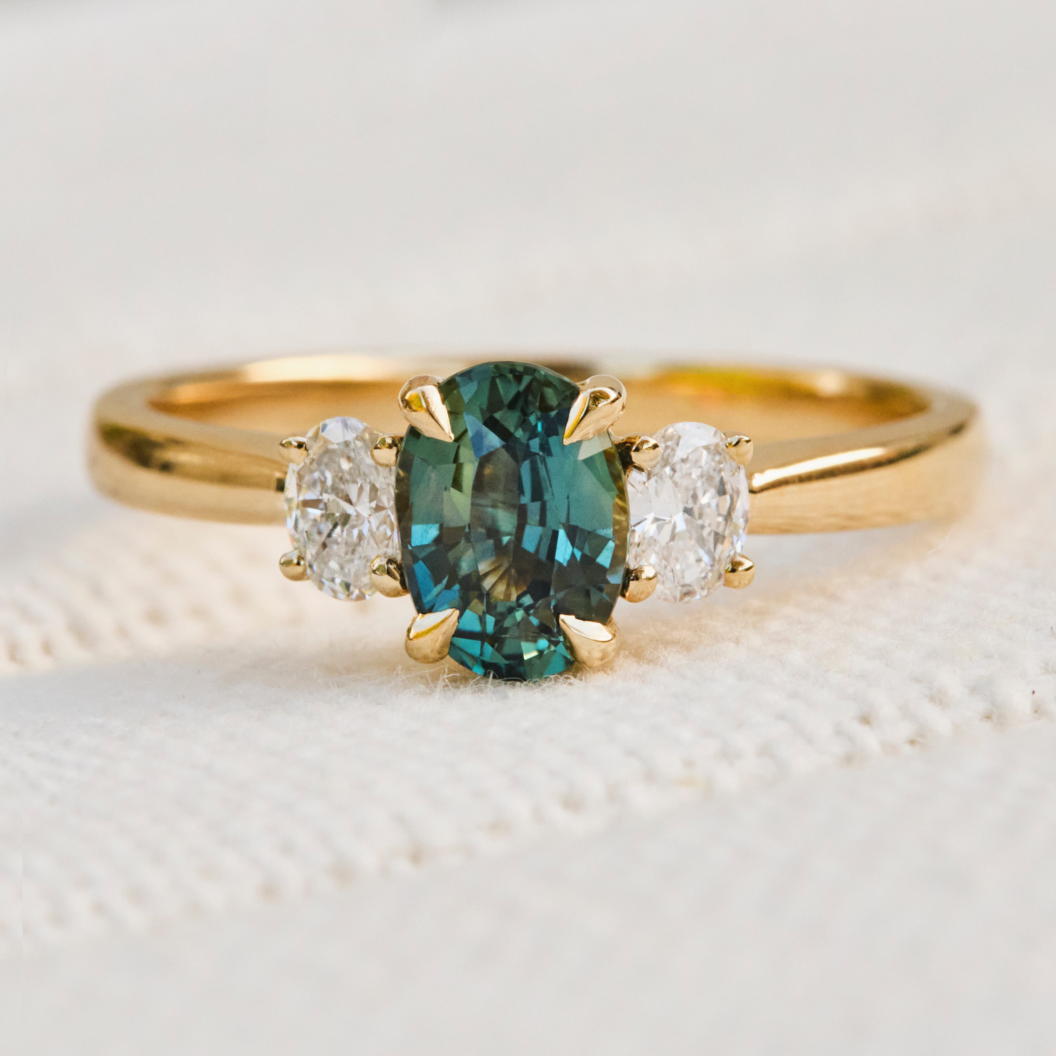 An 18ct yellow gold trilogy ring featuring a central oval-cut teal sapphire flanked by two lab grown diamond side stones.