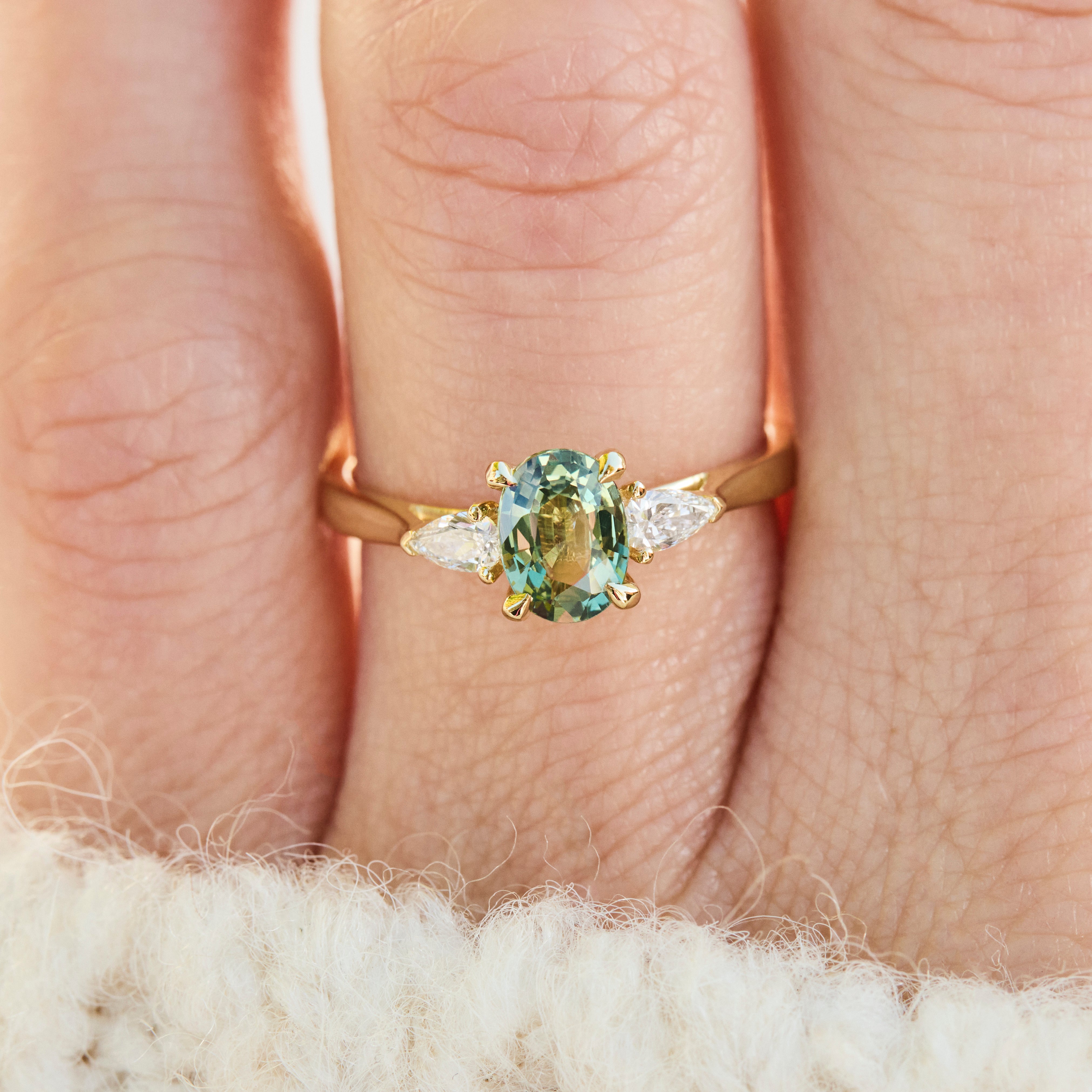 A close-up of an 18ct gold engagement ring with an oval green sapphire centre stone and lab grown pear side diamonds.