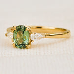 A yellow gold engagement ring featuring an oval-cut green sapphire in a pronged setting  accented by two pear shape lab grown diamonds on either side