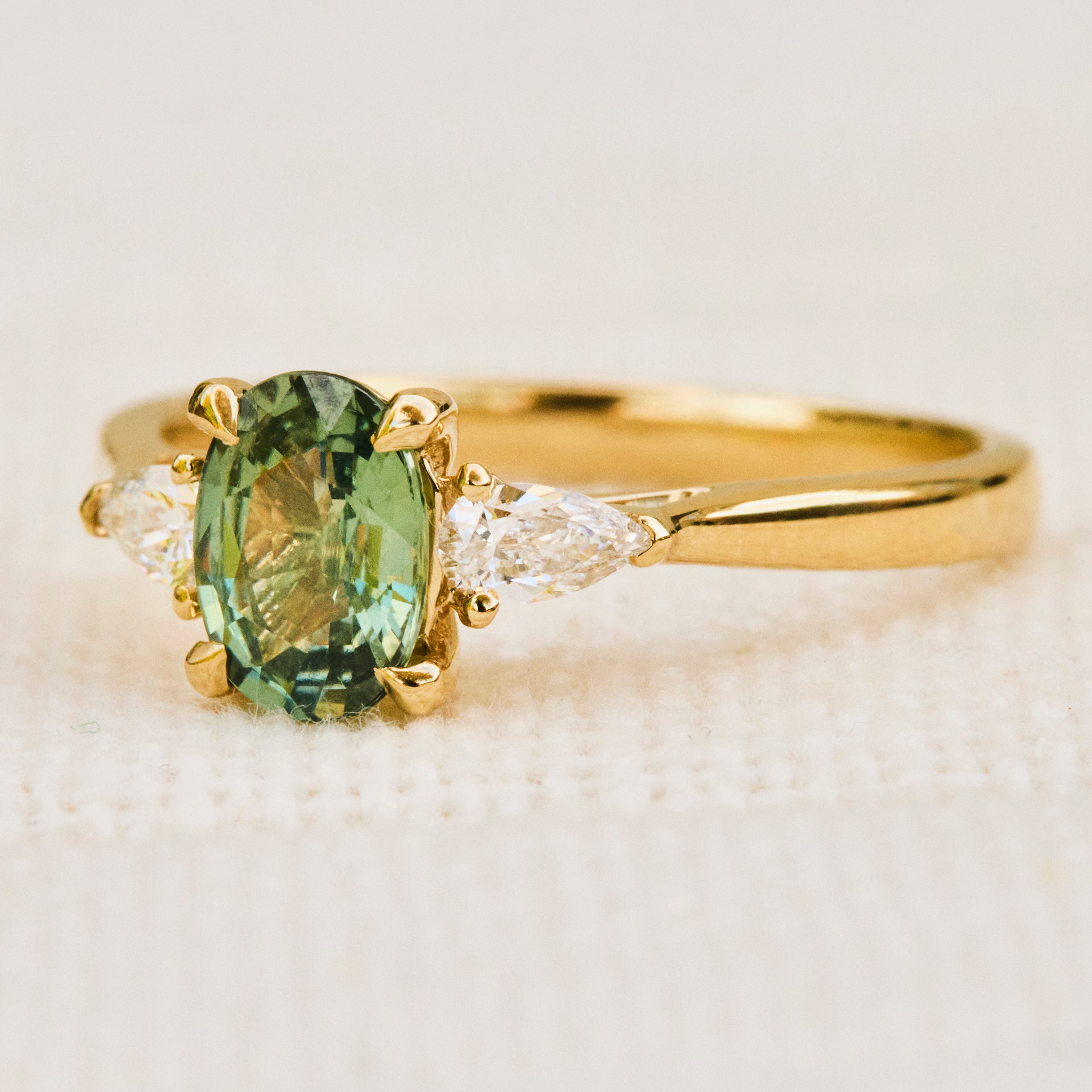 A yellow gold engagement ring featuring an oval-cut green sapphire in a pronged setting  accented by two pear shape lab grown diamonds on either side