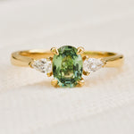 A yellow gold trilogy engagement ring featuring an oval-cut green sapphire in the centre. flanked by two pear shape diamonds.