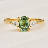 A yellow gold trilogy engagement ring featuring an oval-cut green sapphire in the centre. flanked by two pear shape diamonds.