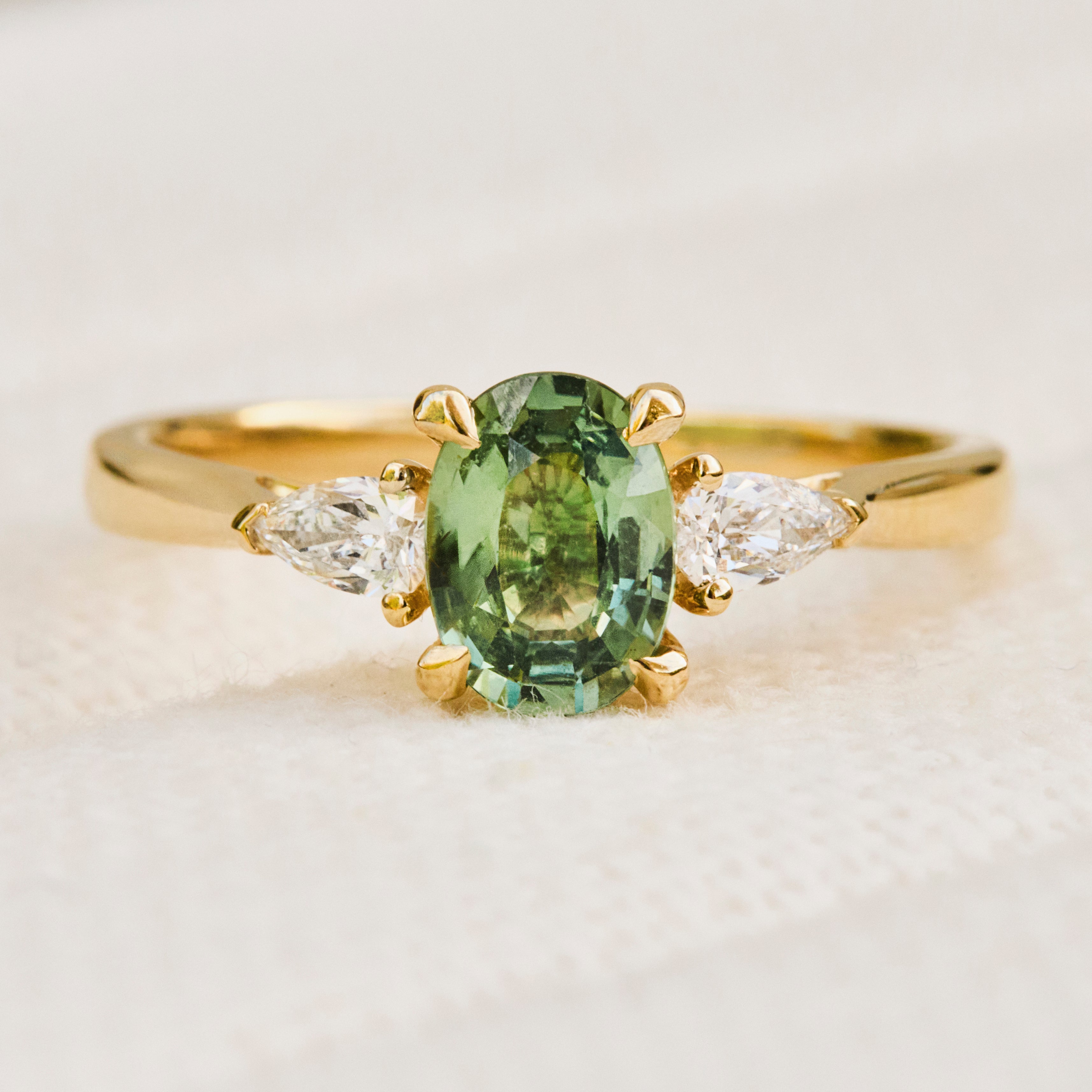A yellow gold trilogy engagement ring featuring an oval-cut green sapphire in the centre. flanked by two pear shape diamonds.