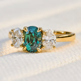An 18ct yellow gold three-stone ring featuring a central oval-teal sapphire flanked by two oval lab grown side stones