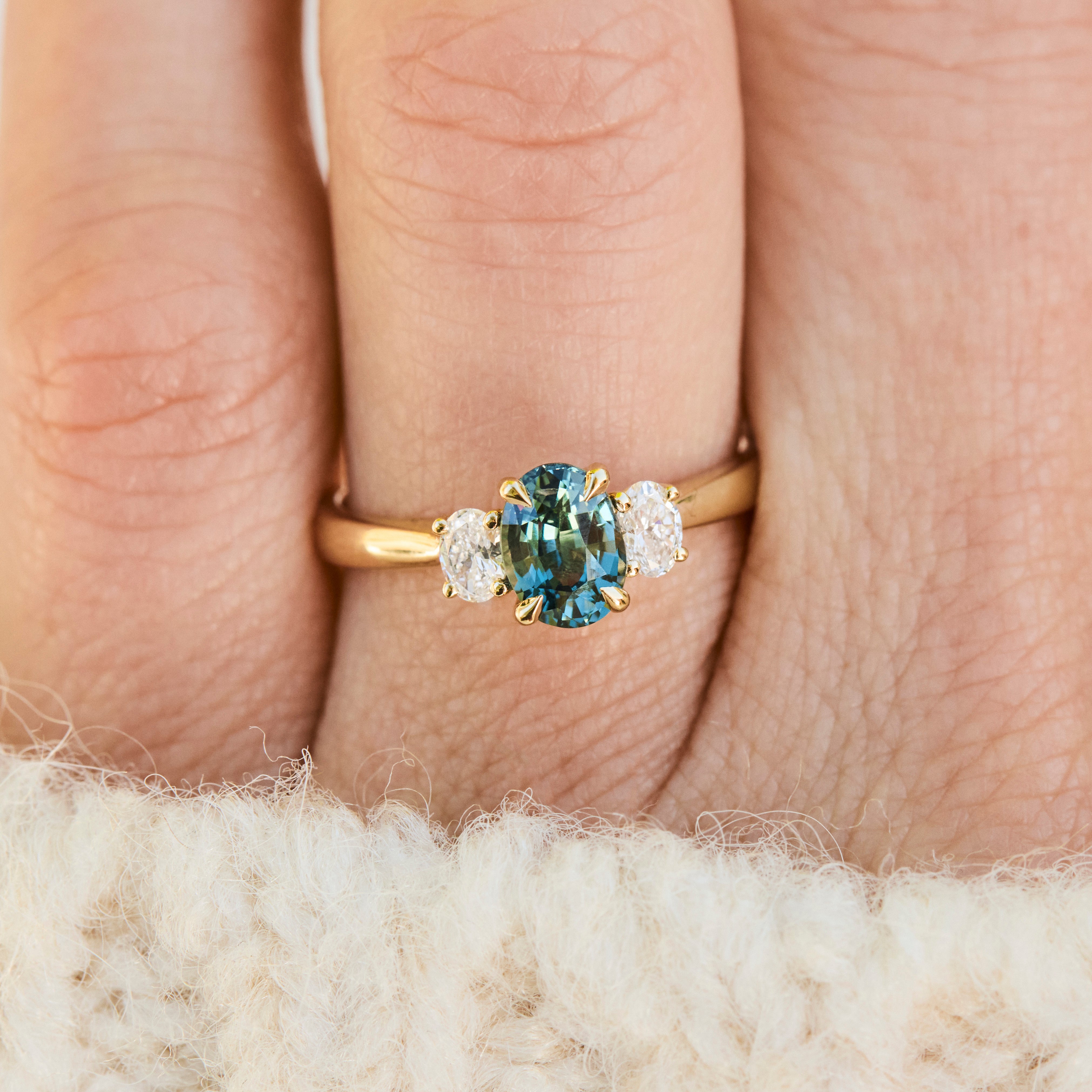 An 18ct  yellow gold engagement ring with a central oval-teal sapphire and two lab grown diamond accent stones.