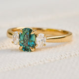 A yellow gold trilogy engagement ring featuring a central oval-cut teal sapphire flanked by two small lab grown side diamonds